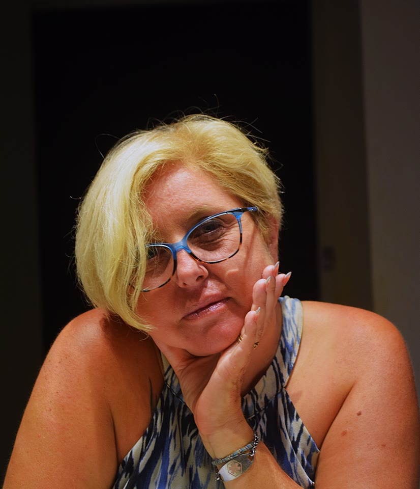 Portrait d’une femme blonde à lunettes, la main posée sur la joue, sur un fond sombre.